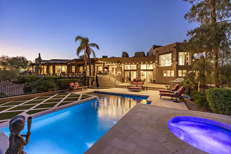Swimming pool and spa glowing at dusk in front of the long, lit residence with palms and patios